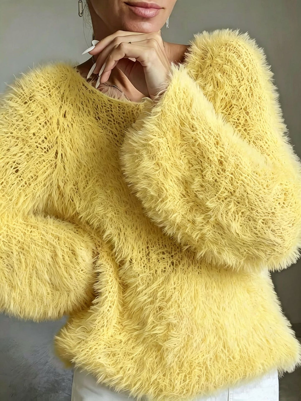 A close-up of a person wearing a fuzzy, textured yellow knit sweater with voluminous long sleeves and a crew neckline, paired with white pants against a neutral gray background.