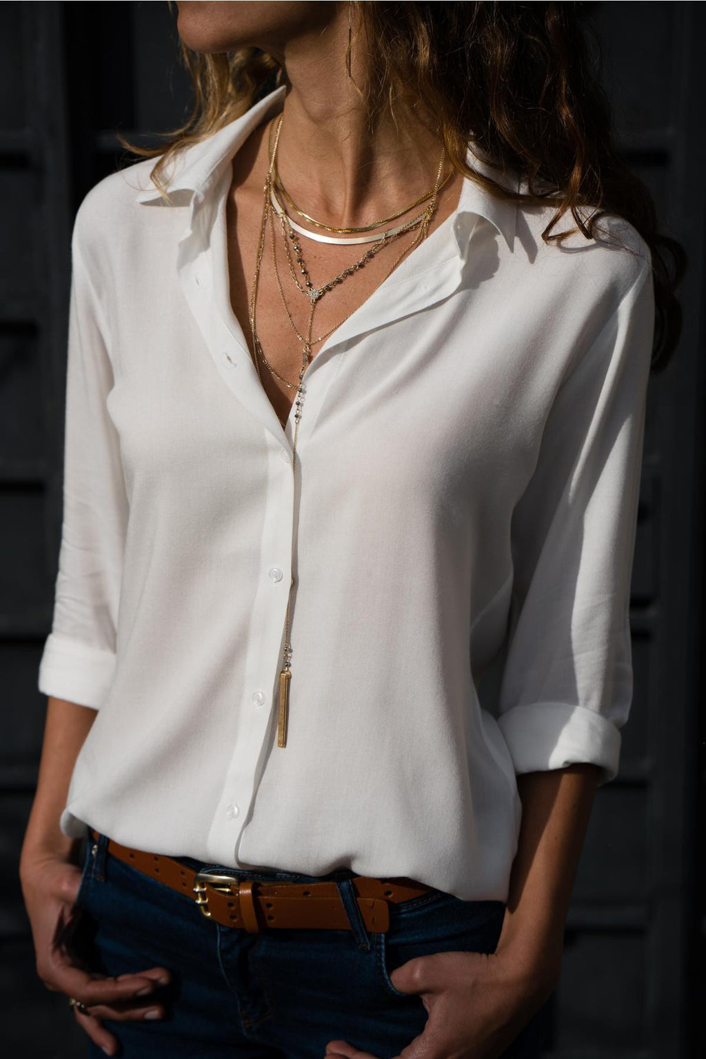 Person wearing a white shirt with rolled-up sleeves and layered necklaces against a dark background