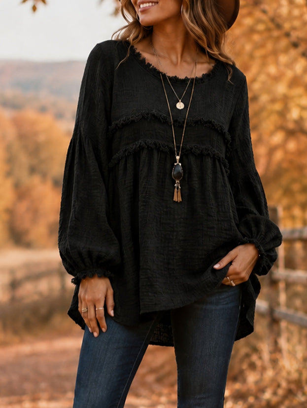 A woman wearing a black long-sleeve blouse with layered ruffle details and a relaxed fit, paired with dark blue jeans, photographed outdoors against a blurred autumn landscape.