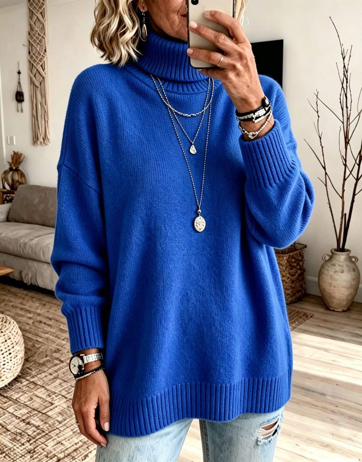 A woman takes a mirror selfie wearing a vibrant royal blue turtleneck sweater, styled with layered silver necklaces, bracelets, and distressed light-wash jeans in a cozy living room setting.