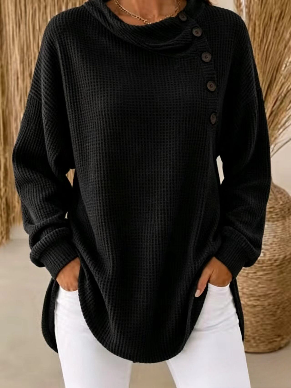 A woman wearing a black waffle-knit cowl-neck sweater with an asymmetrical button closure down one side, paired with white pants.