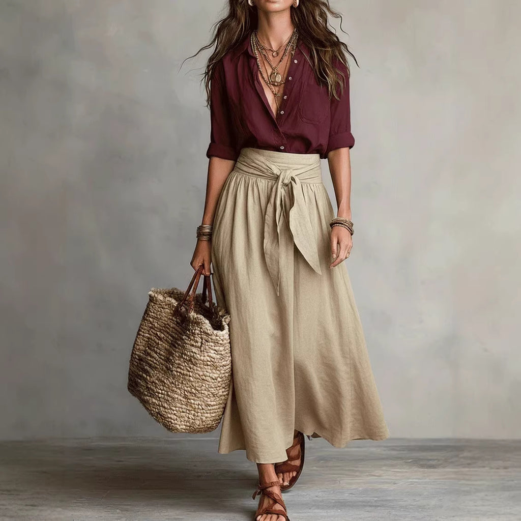 Woman wearing a burgundy shirt and beige skirt holding a woven bag against a neutral background