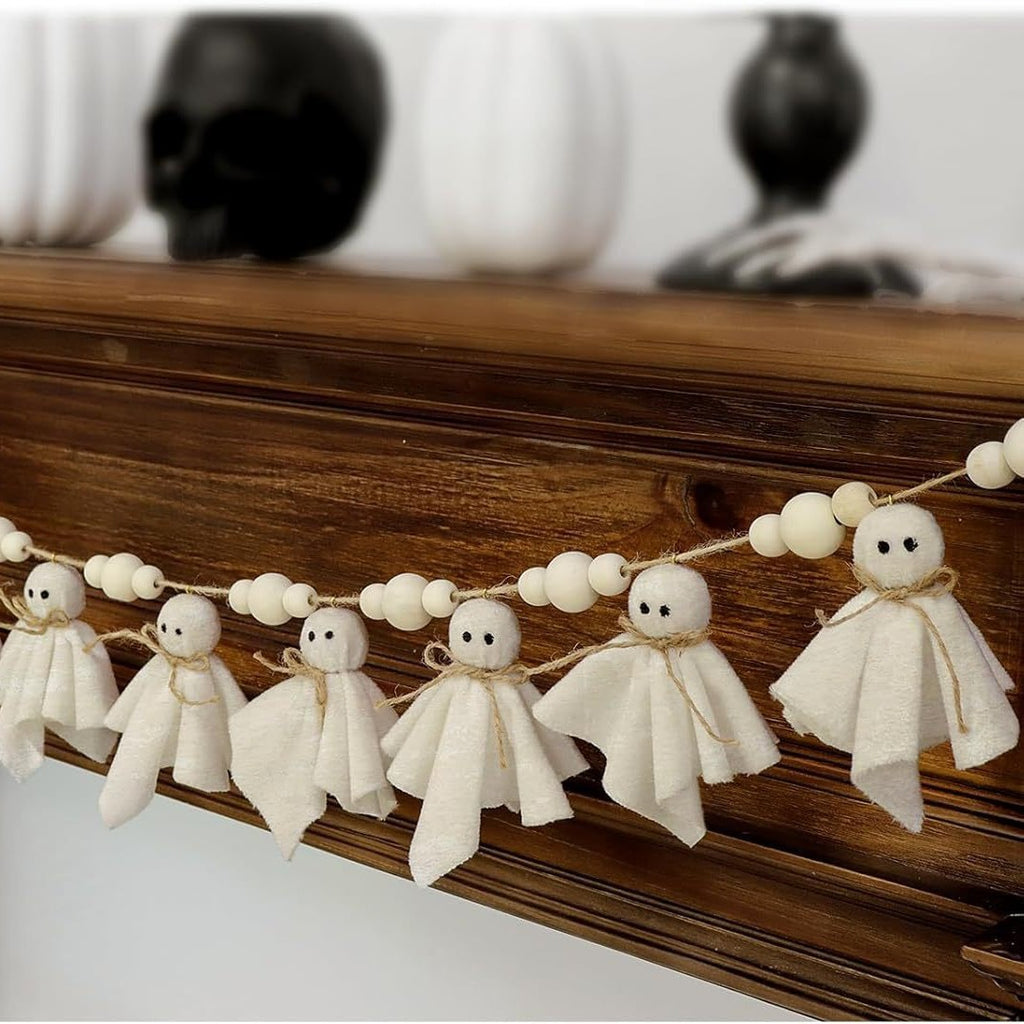 A Halloween garland featuring small felted wool ghost ornaments and white wooden beads strung along a twine, displayed draped over a dark wood mantel with decorative skull vases in the background.