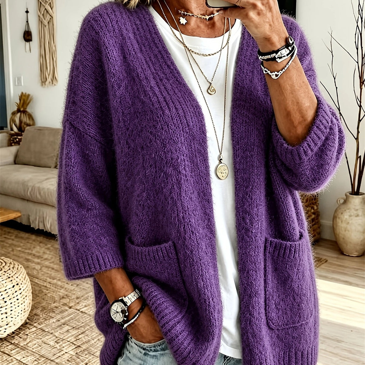 A person wearing a chunky, fuzzy purple open-front cardigan with large front pockets over a white top, photographed indoors in a living room setting.