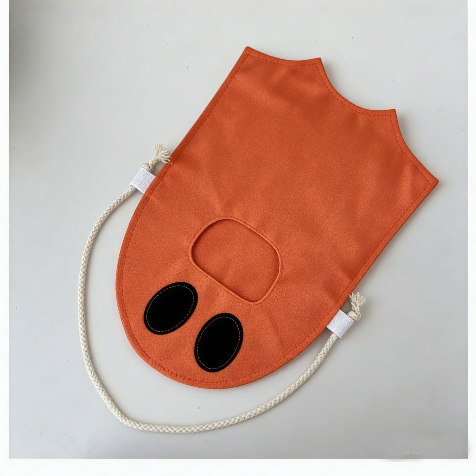 An orange fabric pet hood or face mask with a cutout for the eyes and nose, featuring two black oval patches and a cream-colored braided rope handle, shown on a light gray background.