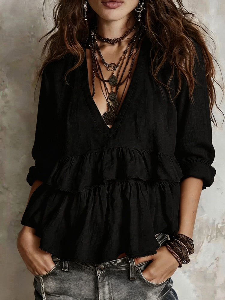 A woman wearing a black bohemian-style long-sleeve blouse with a deep V-neck, layered ruffle details, and rolled cuffs, paired with gray denim jeans and multiple layered necklaces.