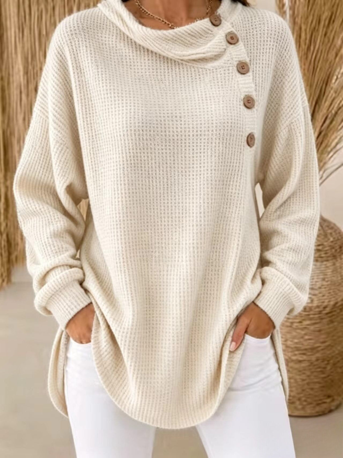 A woman wearing a cream-colored waffle-knit cowl-neck sweater with brown buttons down the side, paired with white pants.