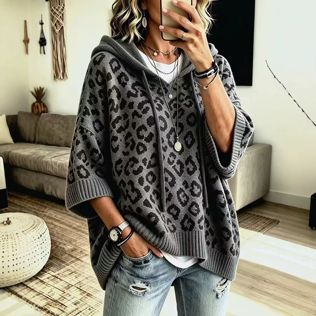 A woman wearing an oversized gray knit hoodie with a black leopard print pattern, layered over a white t-shirt and paired with distressed blue jeans, shown in a mirror selfie in a living room setting.