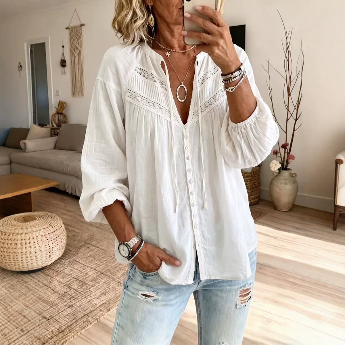 A woman takes a mirror selfie wearing a white long-sleeve button-up blouse with delicate lace embroidery across the chest and light-wash distressed jeans in a bright, modern living room.