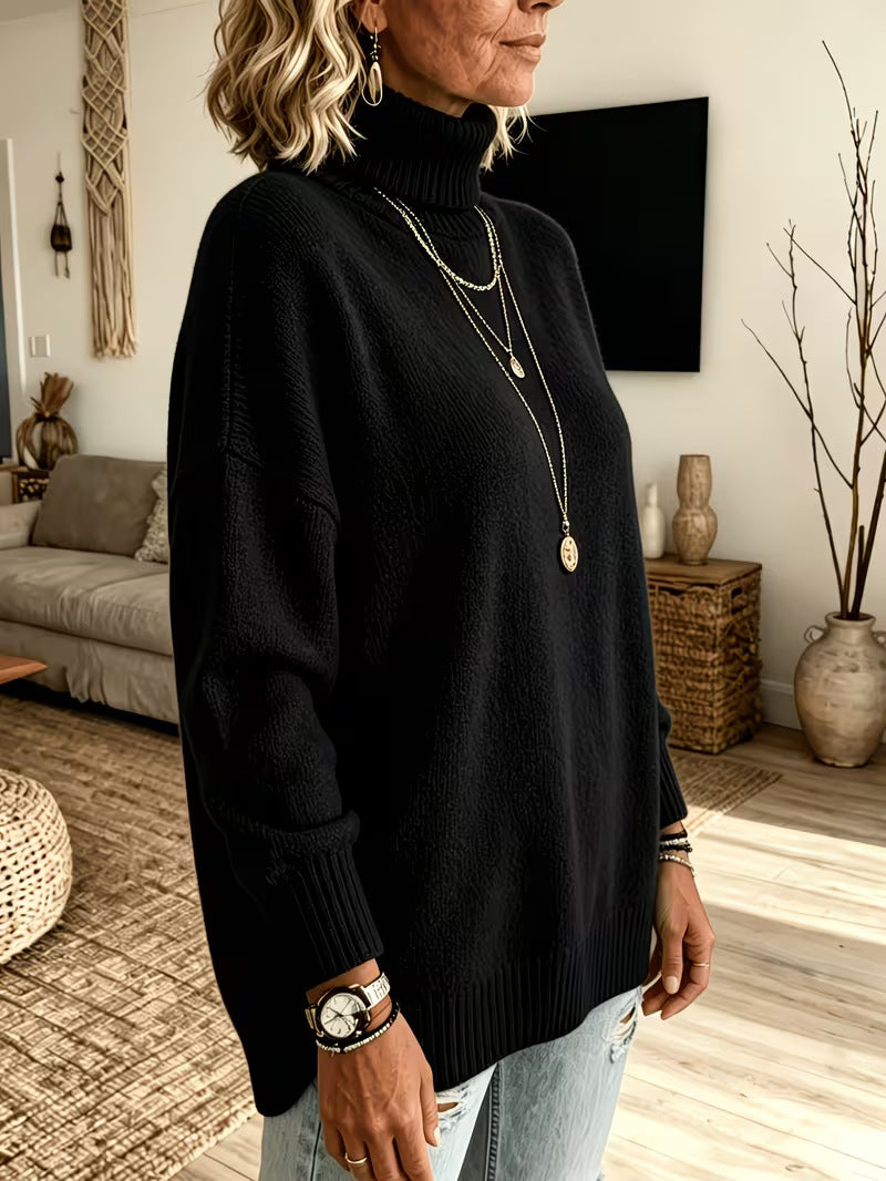 A woman wearing a black ribbed turtleneck sweater, layered with gold necklaces, in a cozy living room setting.