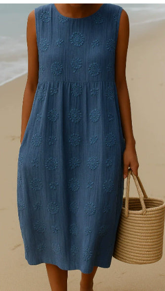 Person wearing a blue dress on a beach holding a woven bag.