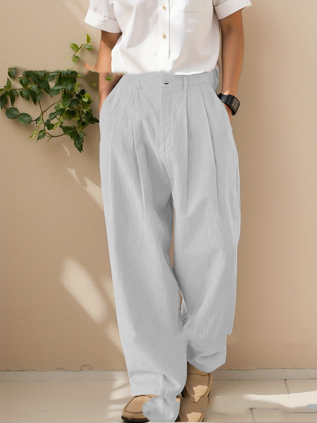 Person wearing light gray pleated pants and a white shirt against a beige wall.