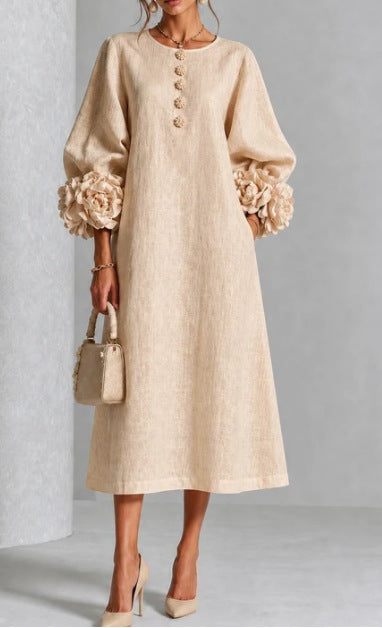 A woman models a beige linen midi dress with voluminous long sleeves featuring large fabric flowers at the cuffs, a round neckline with decorative buttons, and a relaxed A-line silhouette. She is holding a matching beige handbag and wearing beige high heels against a neutral gray studio background.