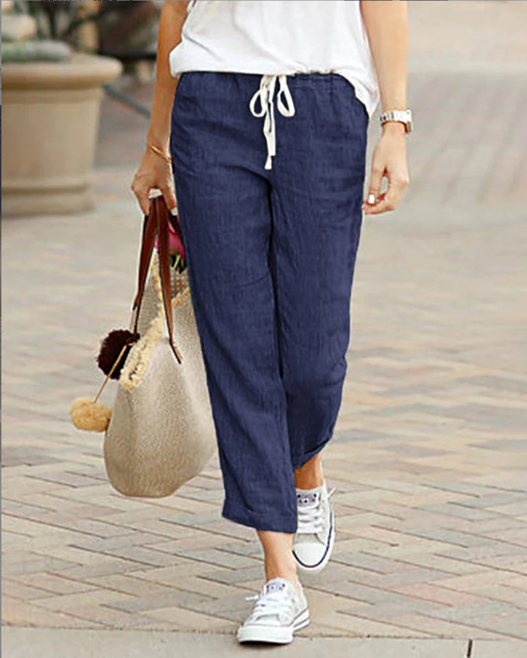 Person wearing navy blue pants with a white top and white sneakers, holding a straw bag on a paved walkway.