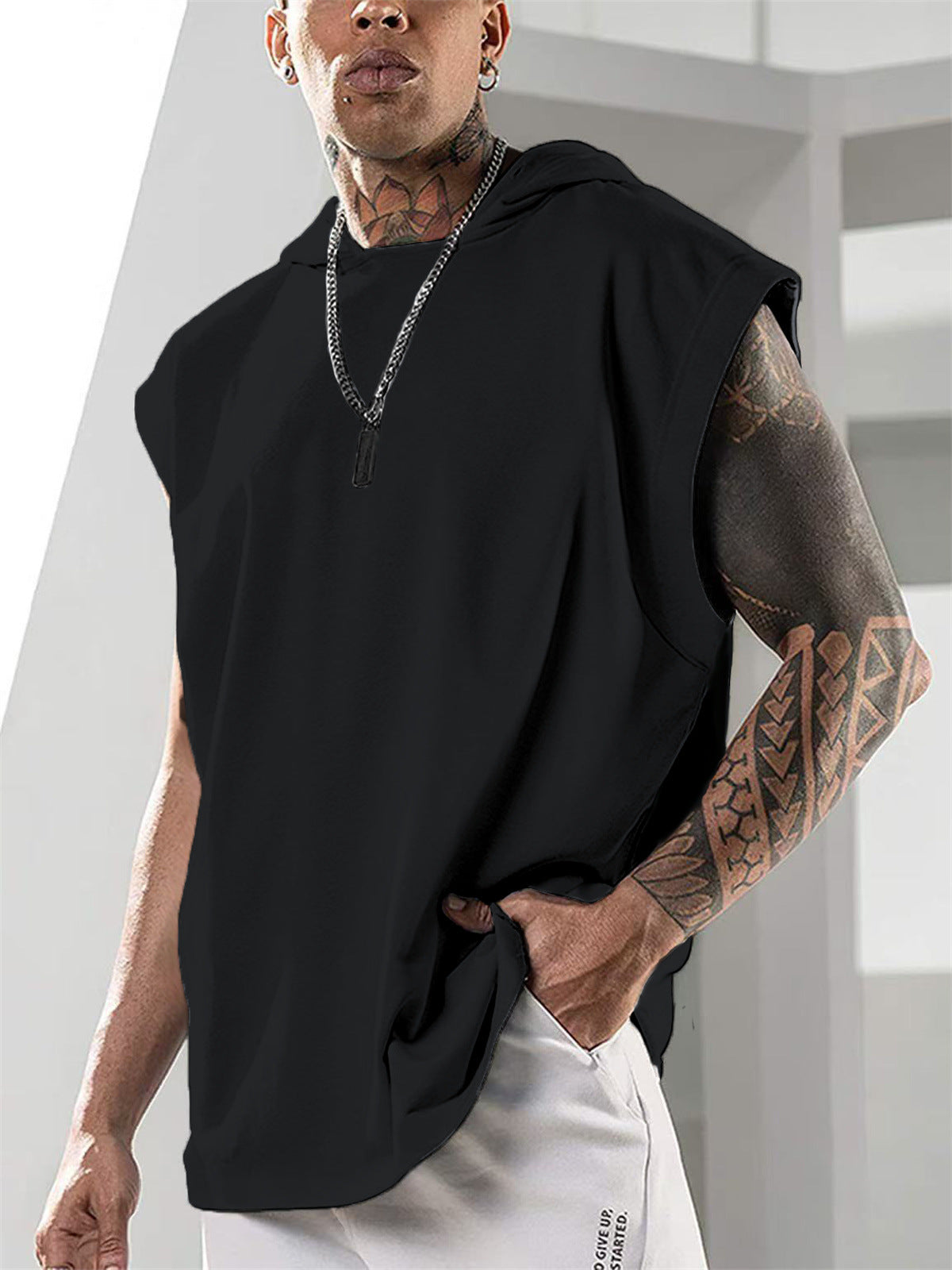 Person wearing a black sleeveless hoodie with tattoos on arms, standing against a light background