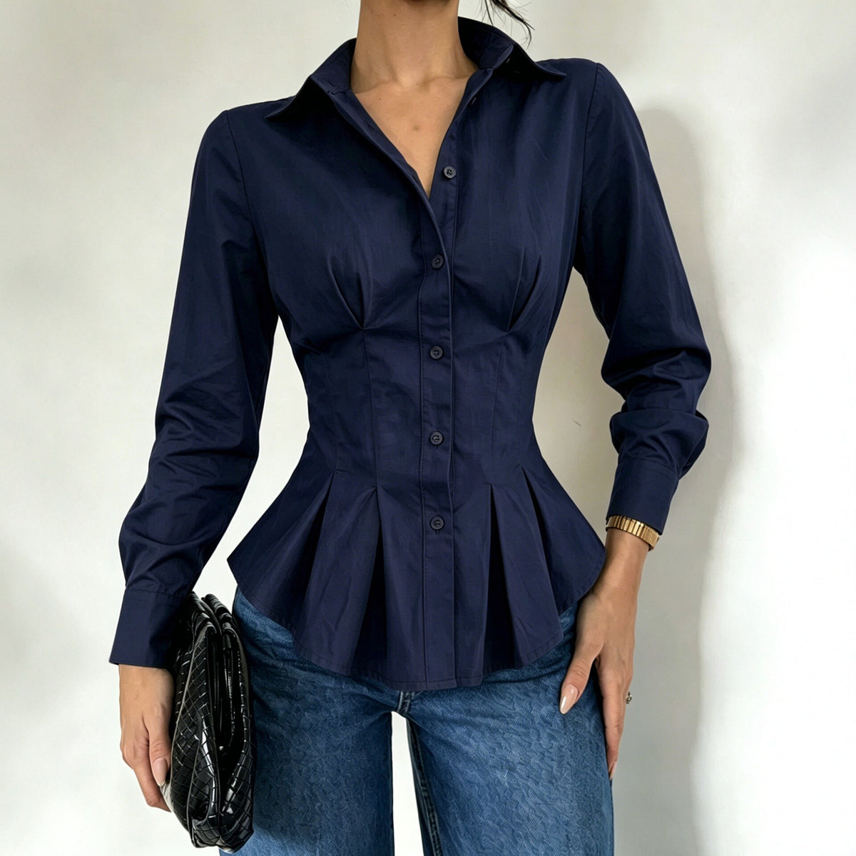 A woman wearing a navy blue long-sleeve button-up blouse with a peplum waist, paired with blue jeans and holding a black clutch bag.