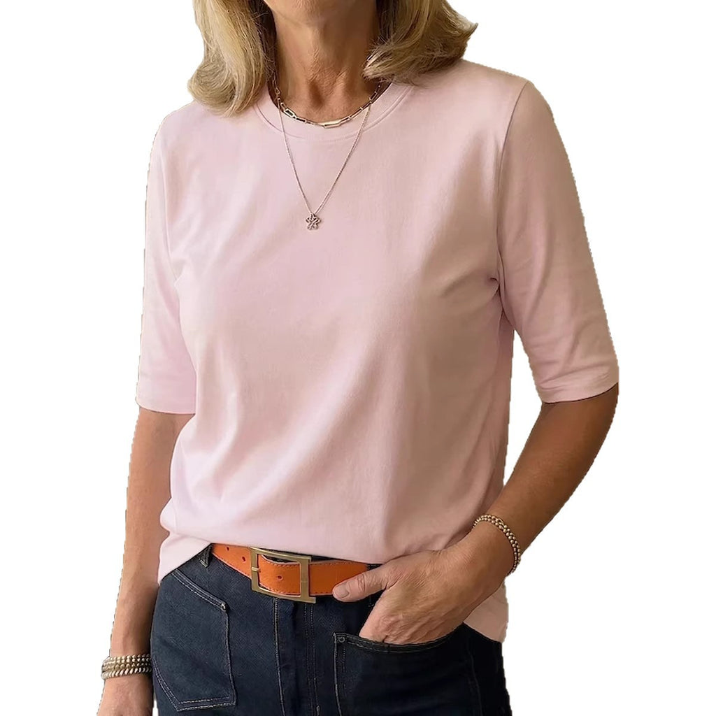 A woman wearing a light pink short-sleeve crewneck top, styled with dark blue jeans, a brown belt, and layered silver necklaces, set against a plain white background.