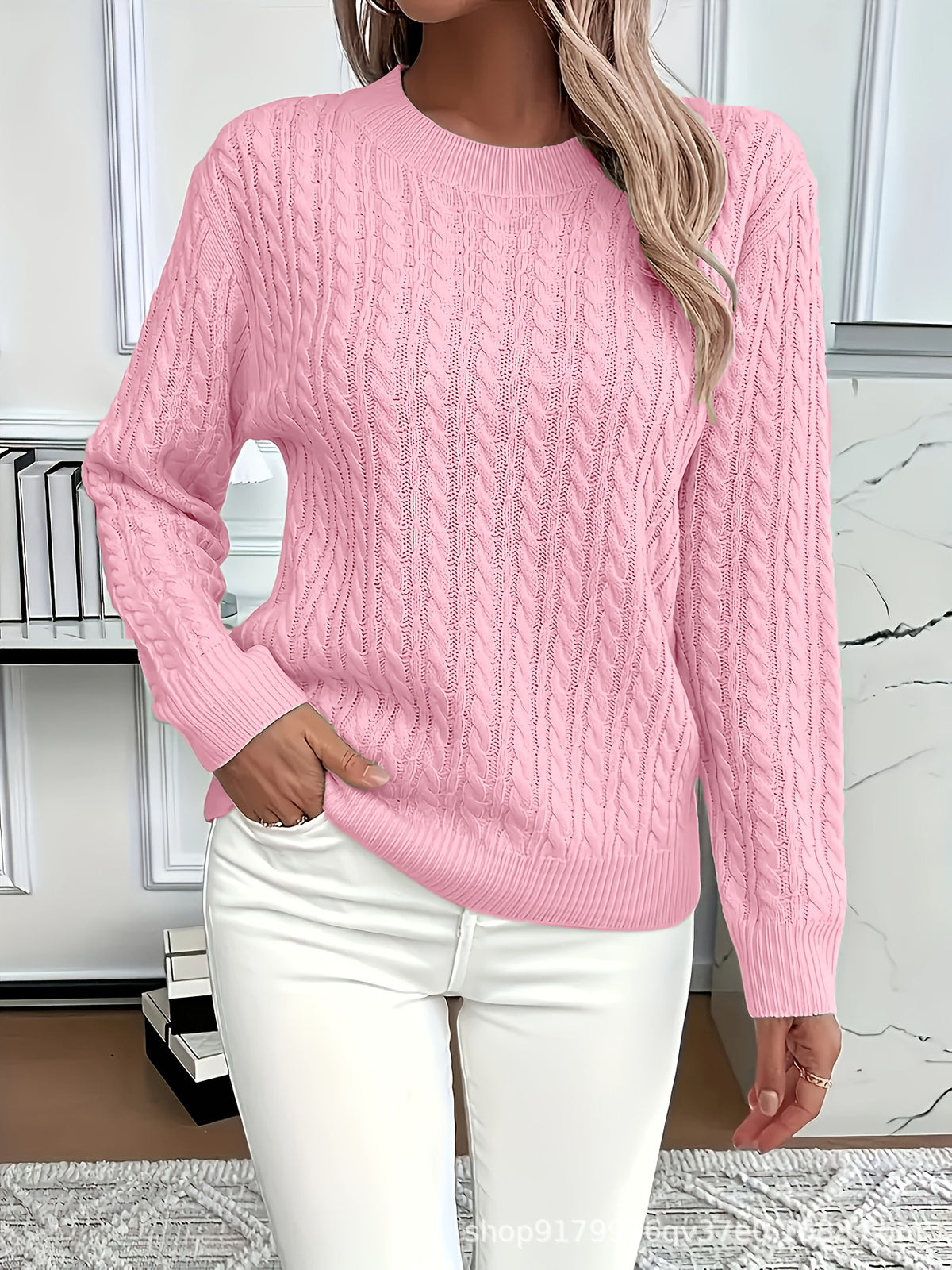 Person wearing a pink cable knit sweater and white pants in an indoor setting.