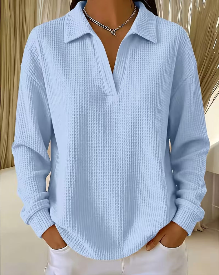 A woman wearing a light blue waffle-knit long-sleeve polo shirt with a split V-neck, paired with white pants and a silver chain necklace.