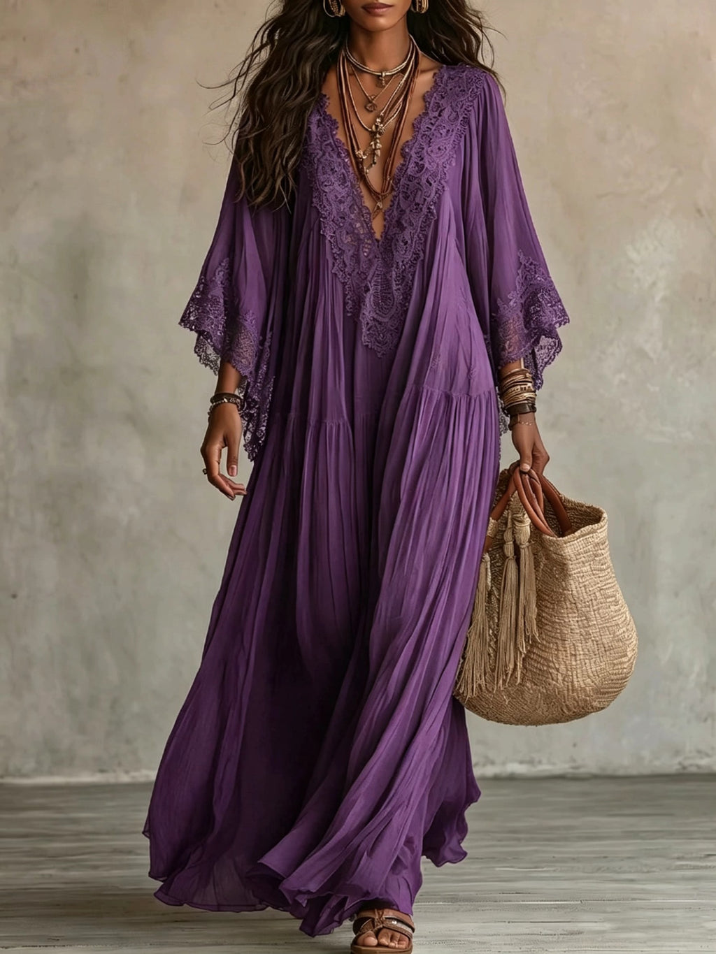 A woman wearing a flowing purple maxi dress with lace trim and wide sleeves, holding a woven straw tote bag against a textured gray wall.