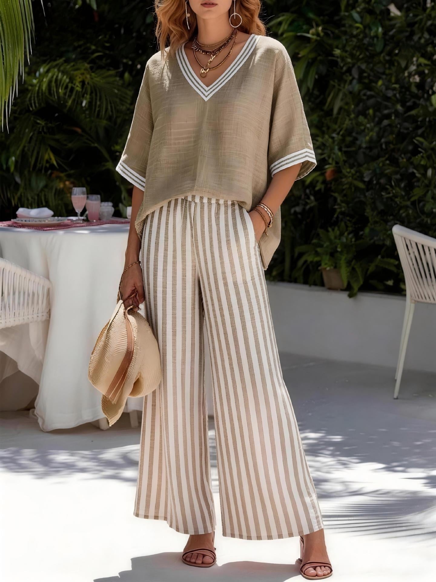 Woman in a beige top and striped pants standing outdoors with greenery and a table in the background.