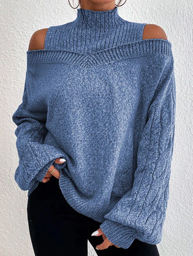 A person wearing a heathered blue cold-shoulder knit sweater with a mock neck and long sleeves, paired with black pants, against a textured off-white wall.