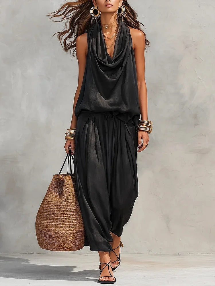 Woman wearing a black sleeveless top and harem pants, holding a woven bag against a neutral background.