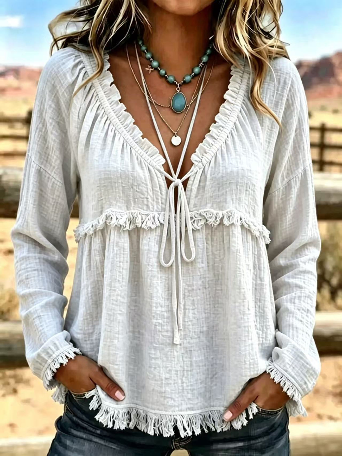 A woman wearing a white long-sleeve bohemian-style blouse with ruffled trim, a tie-front neckline, and fringe details, paired with dark jeans and layered necklaces, set against a blurred outdoor desert background.