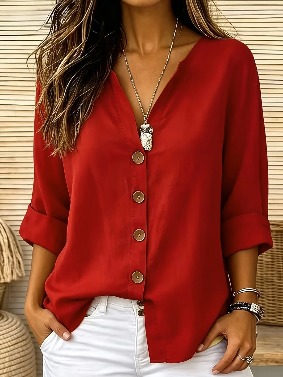 A woman wearing a red button-up blouse with rolled sleeves and white pants, accessorized with a silver pendant necklace and bracelets, set against a background of beige wicker baskets.