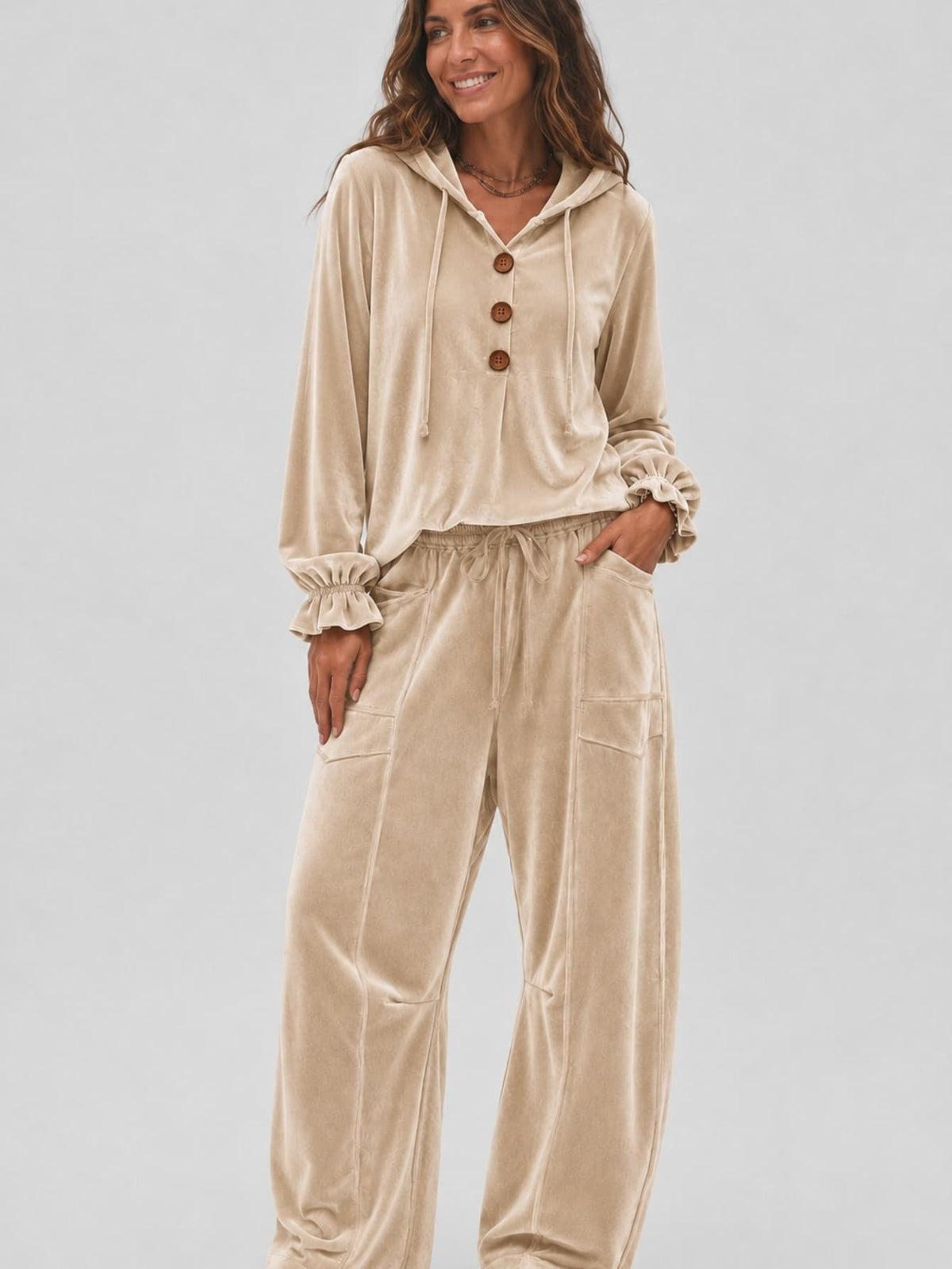 A woman modeling a beige velvet two-piece loungewear set consisting of a long-sleeve hooded top with three brown buttons and matching wide-leg pants, shot against a light gray studio background.
