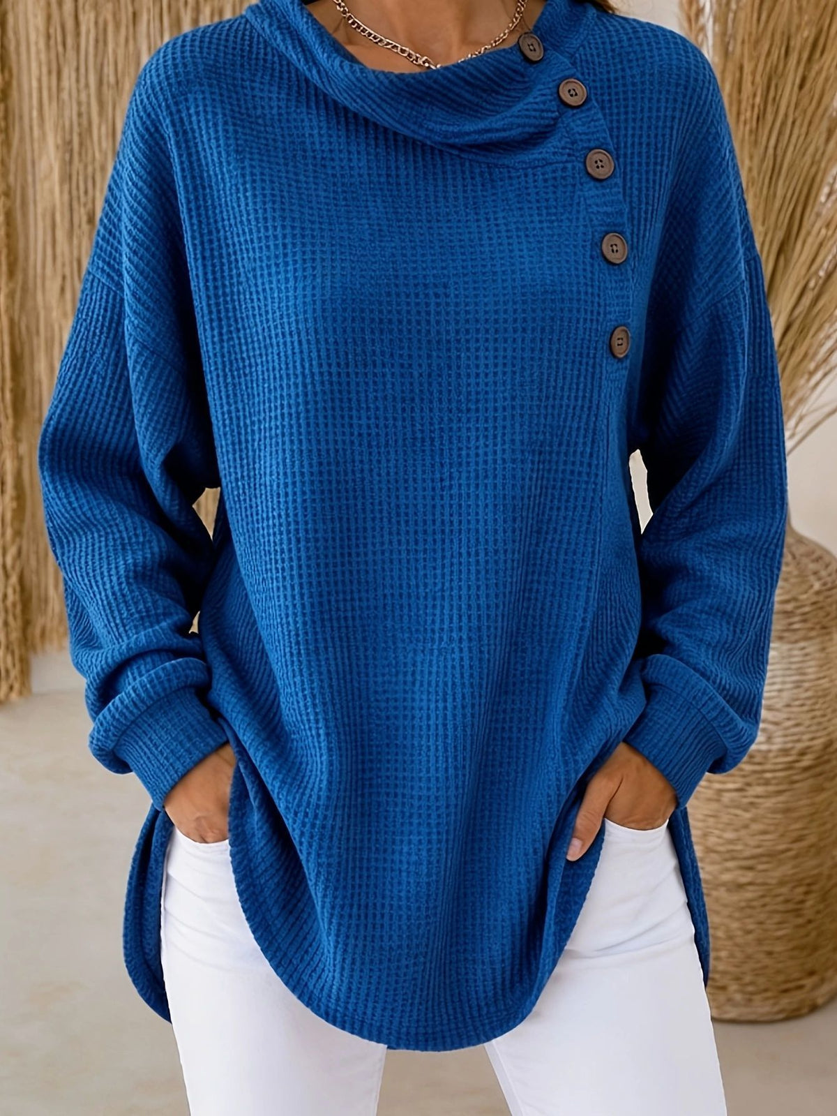 A person wearing a blue waffle-knit cowl-neck sweater with brown buttons down the side, paired with white pants, standing indoors against a neutral woven background.