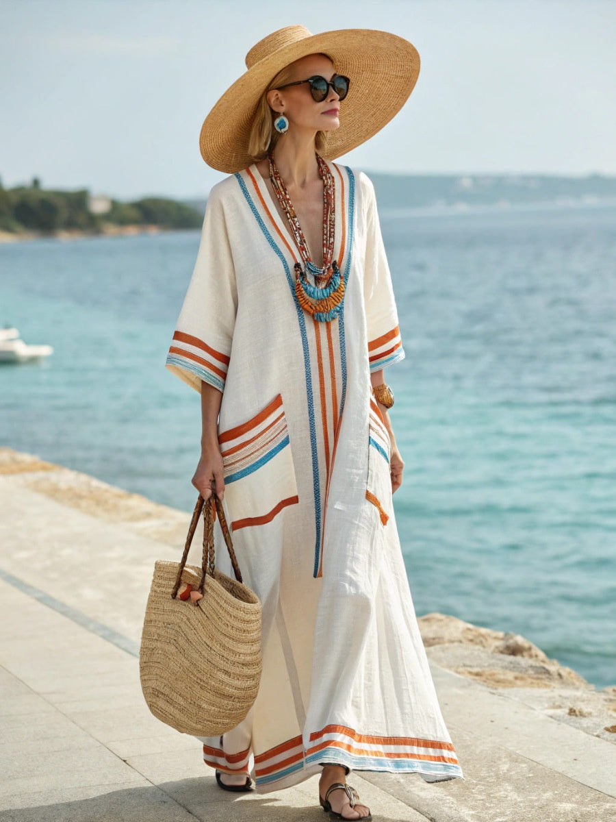 Woman in a long, light-colored dress with colorful stripes, straw hat, and sunglasses by the sea.