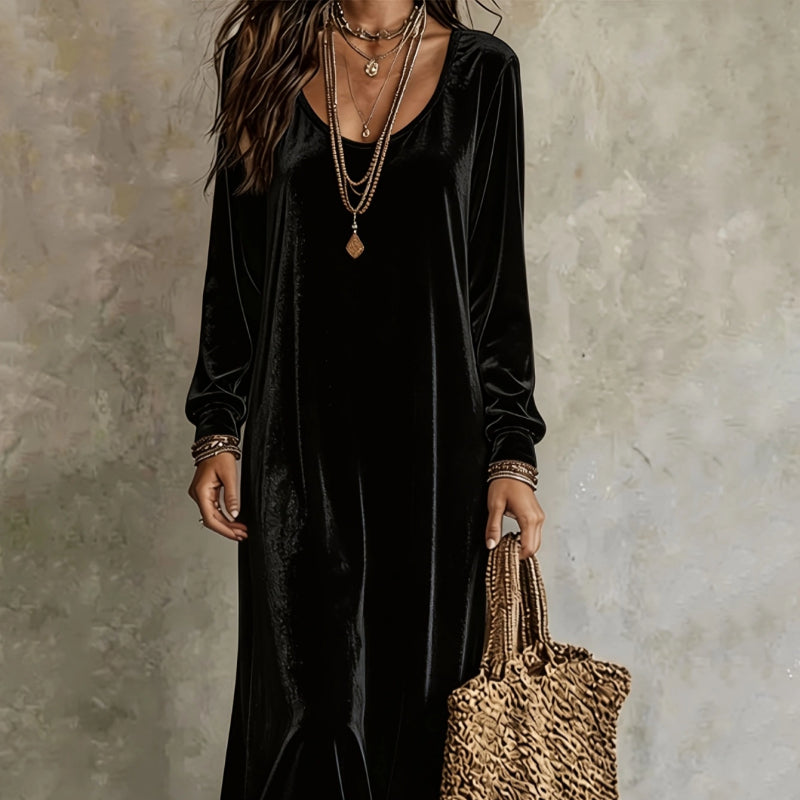 A woman wearing a long black velvet dress with long sleeves and a scoop neckline, accessorized with layered gold necklaces, bracelets, and a woven tan tote bag, set against a textured gray wall.