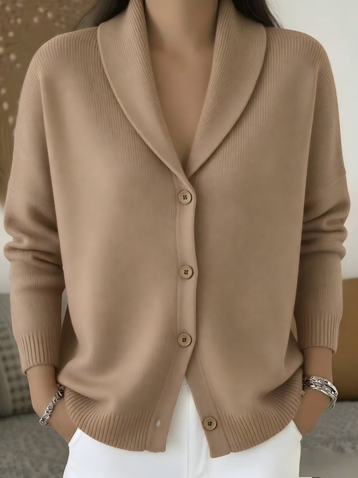 A woman wearing a beige ribbed knit cardigan sweater with a shawl collar and front buttons, paired with white pants.