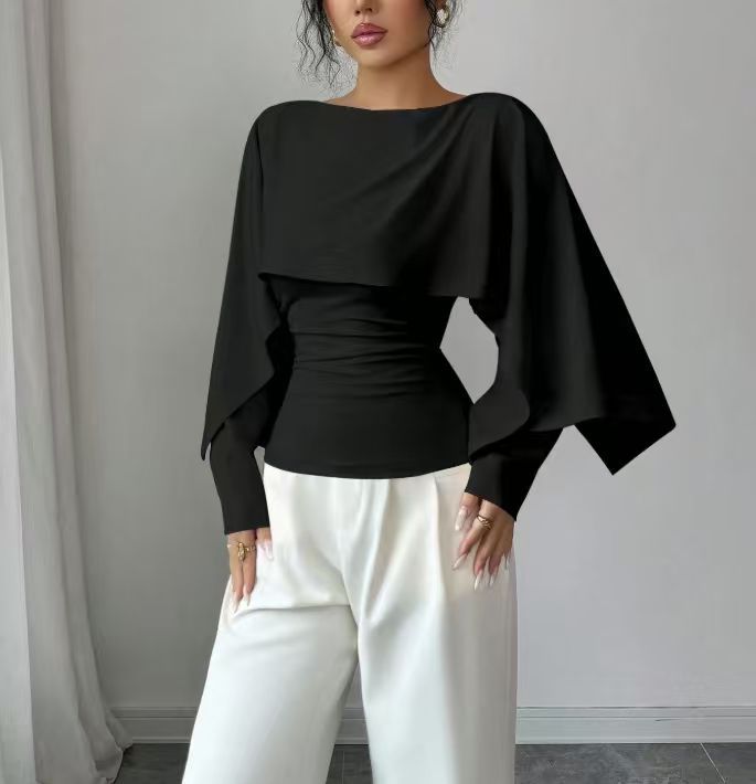A woman wearing a black long-sleeve top with a draped, layered overlay and white wide-leg pants, standing against a light gray wall.
