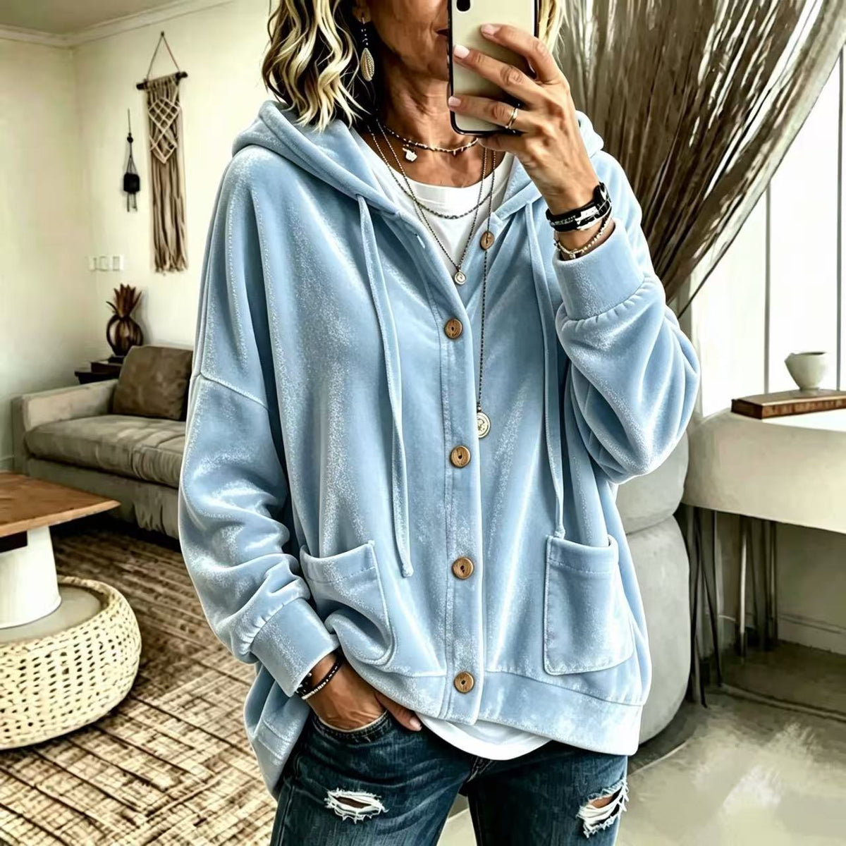 A woman takes a mirror selfie wearing a light blue velour hoodie with brown buttons, layered over a white t-shirt and paired with distressed blue jeans in a modern living room setting.