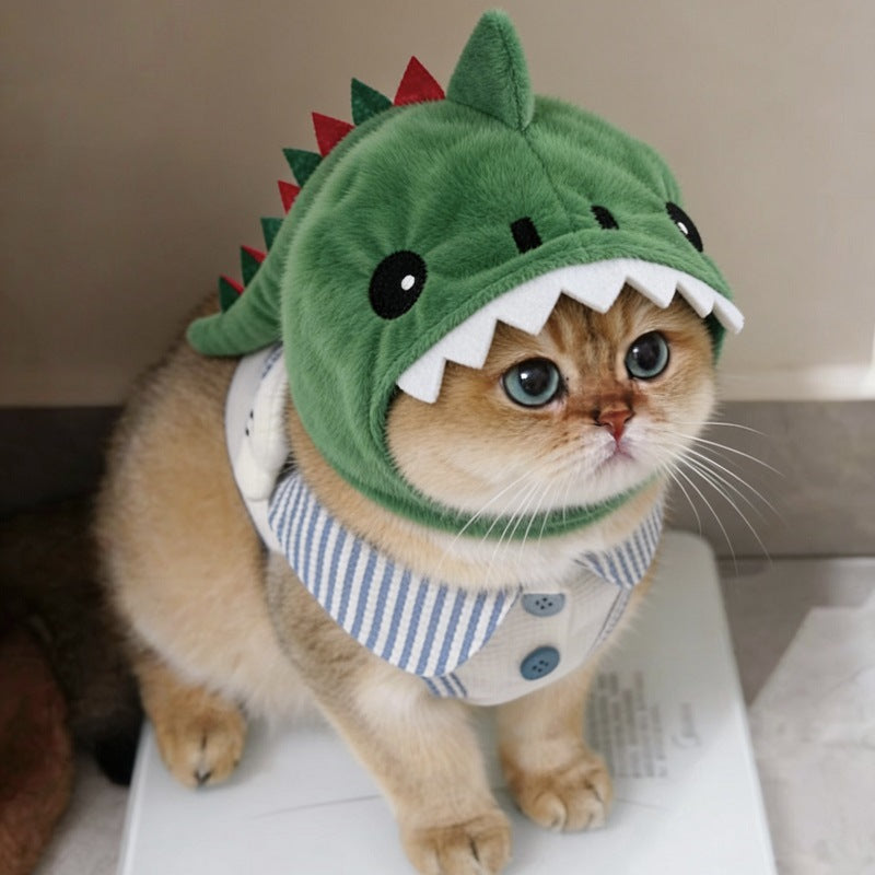 A golden cat wearing a plush green dinosaur costume hood with white teeth and a blue-and-white striped shirt, standing on a white surface indoors.