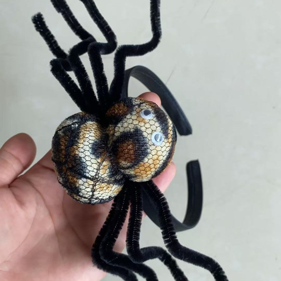 A handmade spider costume accessory held in a hand, featuring two mesh-covered spheres with a tan and black snakeskin pattern, black pipe cleaner legs, and a black elastic headband.