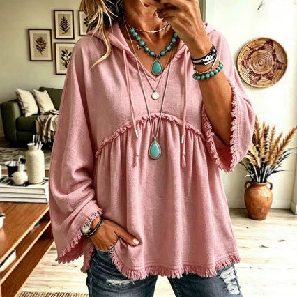 A woman wearing a dusty pink hooded tunic top with fringe trim and wide bell sleeves, paired with distressed blue jeans and layered turquoise jewelry, standing in a living room setting.