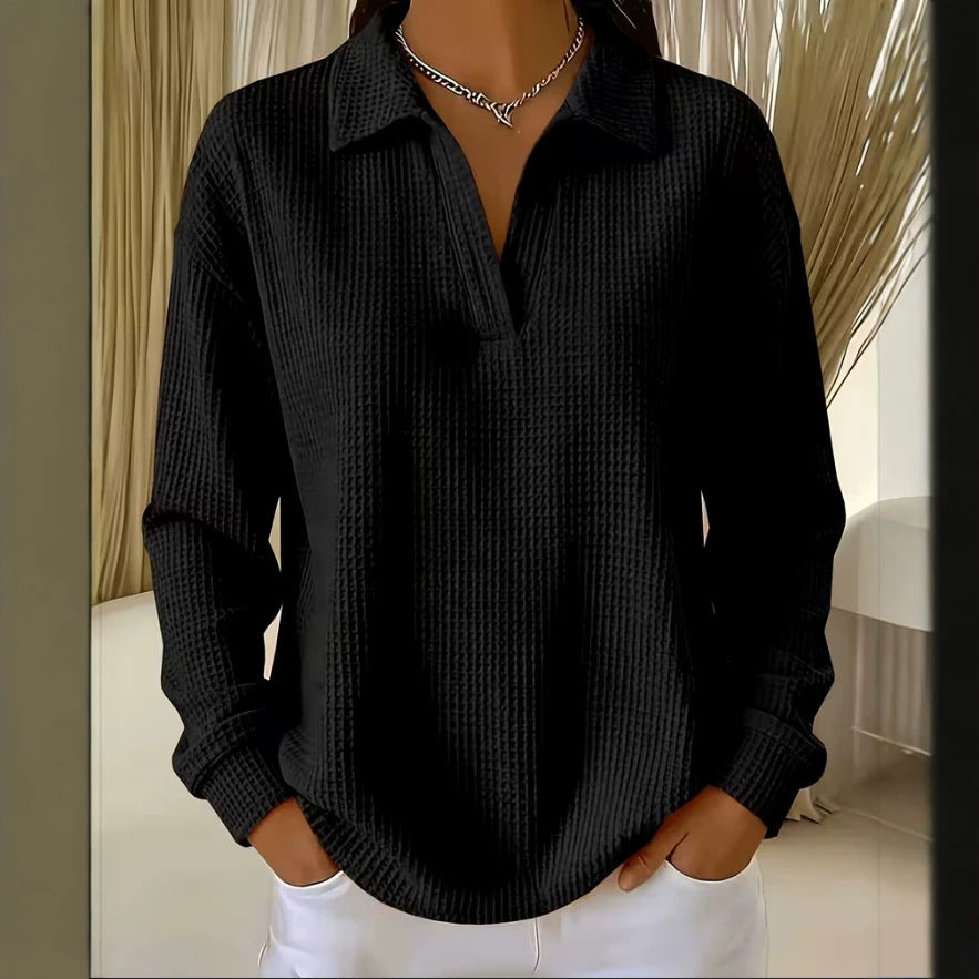 A woman wearing a black long-sleeve collared knit top with a textured waffle pattern, paired with white pants and a silver chain necklace, shown in an indoor setting.