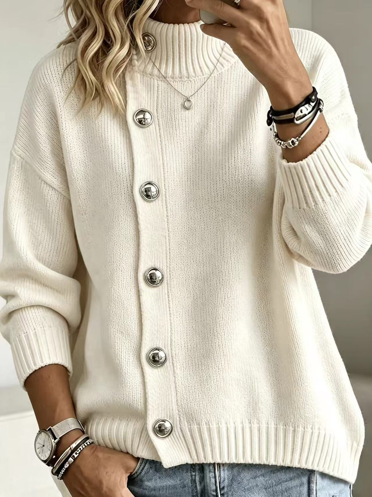 A woman wearing a cream-colored ribbed knit cardigan sweater with large silver buttons, paired with blue jeans and accessorized with bracelets and a necklace.