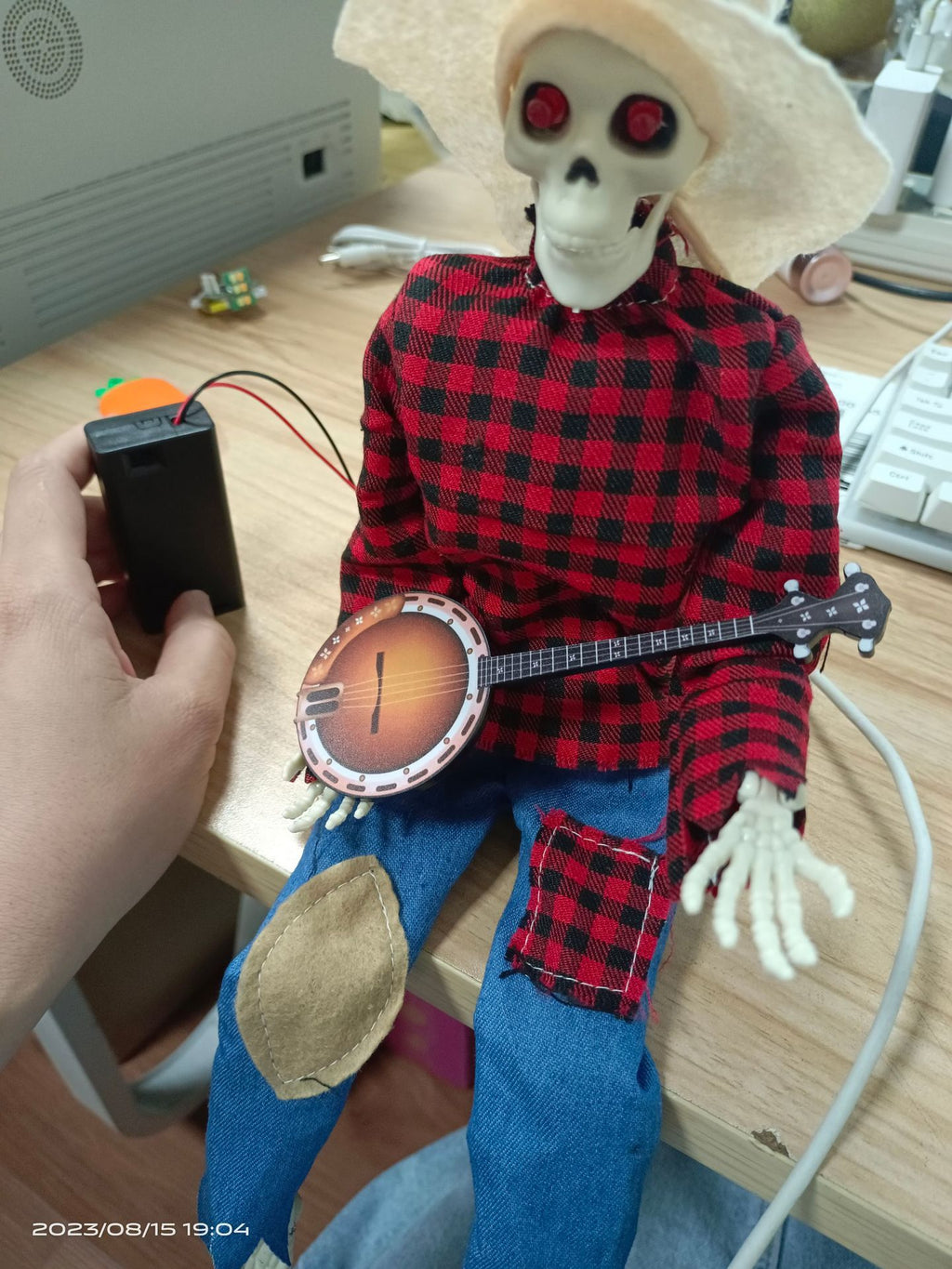 A skeleton doll wearing a beige cowboy hat, a red and black plaid long-sleeve shirt, and blue jeans with patch details, holding a banjo. A human hand is holding a black power battery connected to the doll.