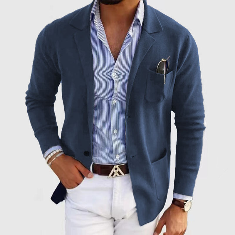 Man wearing a blue blazer over a striped shirt with white pants on a light gray background
