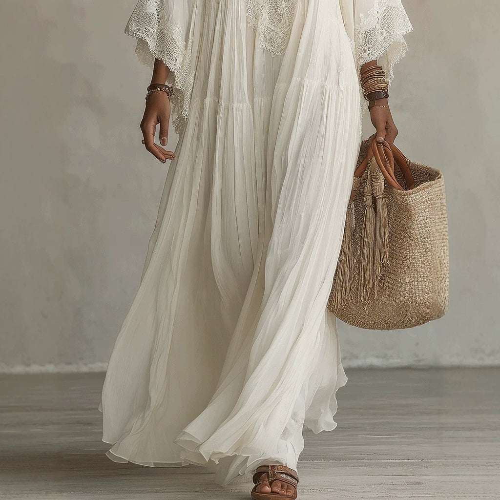 A woman wearing a flowing white maxi dress with lace flutter sleeves and brown strappy sandals, holding a woven straw tote bag with tassel straps against a neutral studio backdrop.
