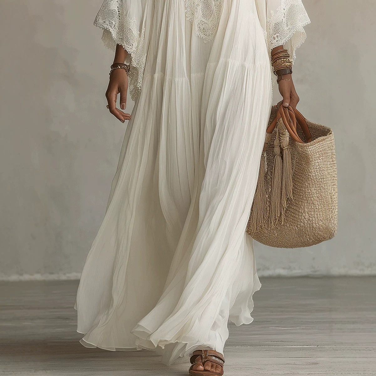 A woman wearing a flowing white maxi dress with lace flutter sleeves and brown strappy sandals, holding a woven straw tote bag with tassel straps against a neutral studio backdrop.