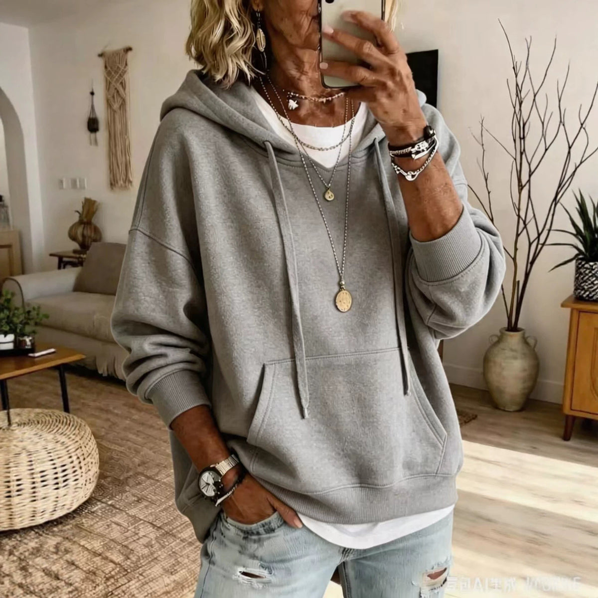 A woman takes a mirror selfie in a cozy living room, wearing a light gray oversized pullover hoodie over a white t-shirt and light-wash distressed jeans.