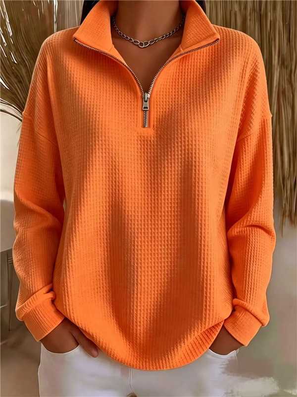 A person wearing a bright orange waffle-knit quarter-zip pullover sweater with a stand collar and white pants, shown from the neck down in an indoor setting.