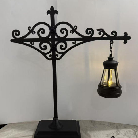Black wrought iron decorative stand with an ornate scrollwork top and a hanging lantern-style light on a marble-like surface against a plain light background.