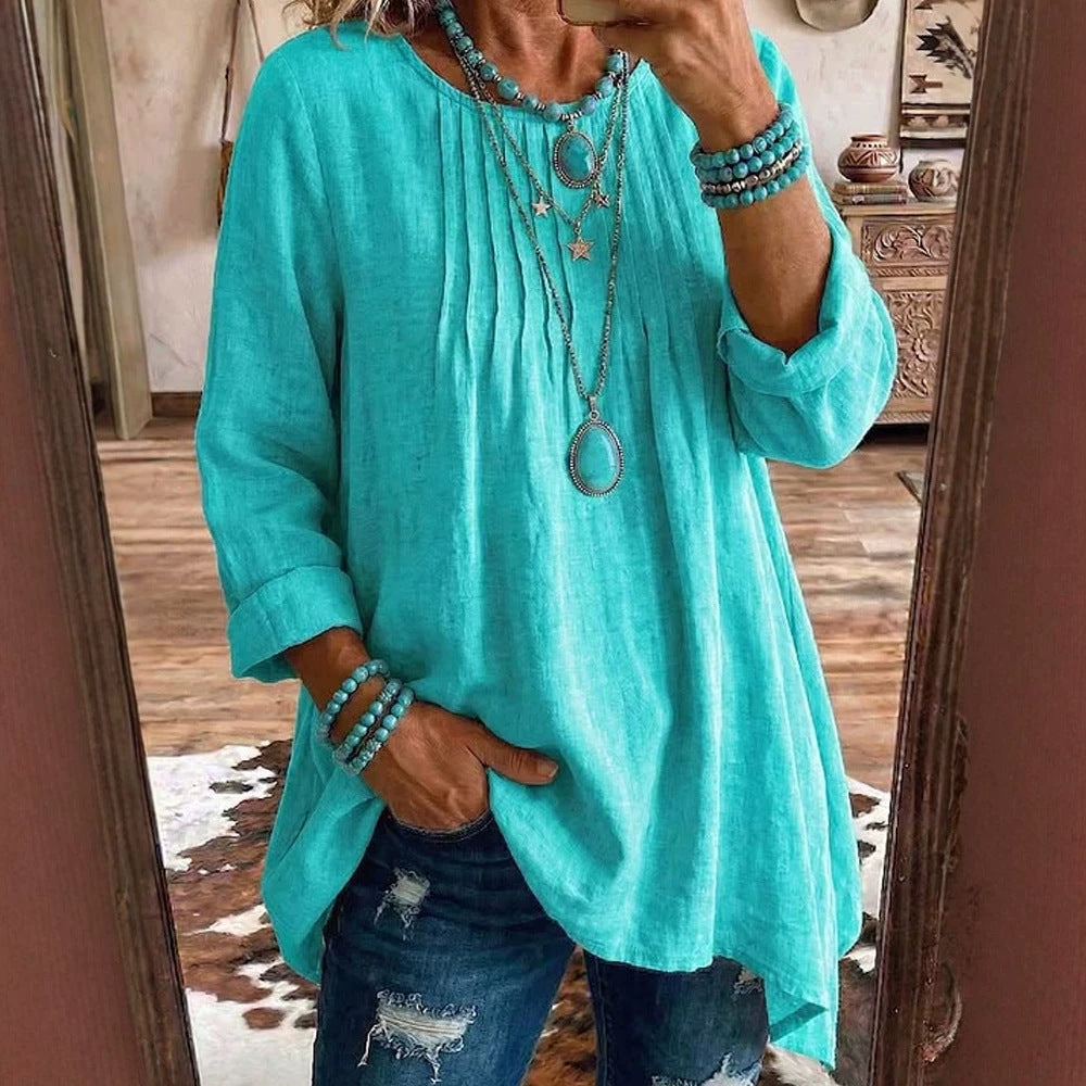 A woman takes a mirror selfie wearing a loose, turquoise long-sleeve linen top with pleated detailing on the chest, paired with distressed blue jeans and layered turquoise and silver jewelry.