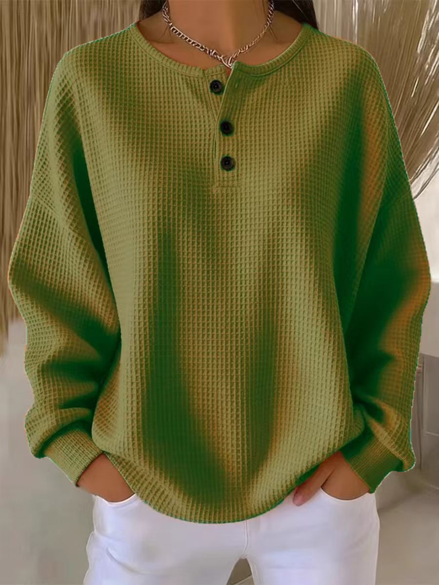 A person wearing an olive green waffle-knit long-sleeve henley top with three black buttons, paired with white pants.
